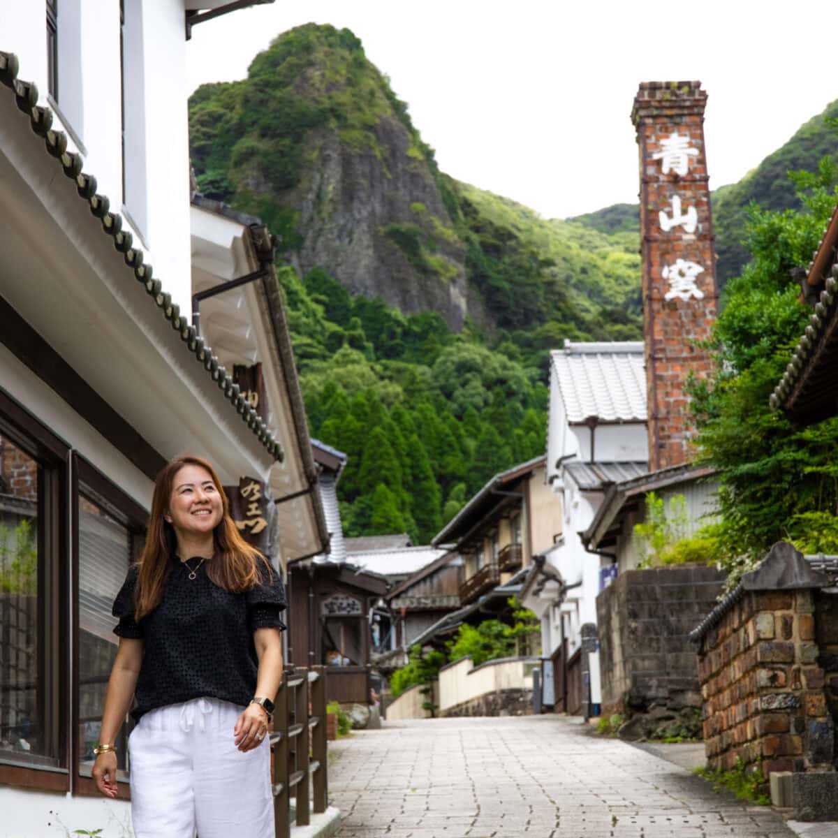 Picturesque Japanese mountain village with traditional architecture and lush greenery, featuring a smiling woman walking along a cobblestone street, capturing the charm of rural Japan and cultural heritage.