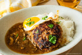 A plate with a serving of white rice topped with a fried egg, a browned hamburger patty, brown mushroom gravy, and garnished with fresh herbs. A drink and a yellow towel are in the background.