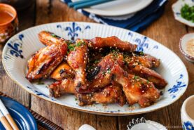 A platter of glazed chicken wings garnished with chopped green herbs and sesame seeds, served on a decorative oval plate on a wooden table.