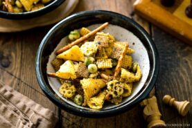 A bowl of snack mix featuring Chex cereal, pretzel sticks, rice crackers, and green wasabi peas sits on a wooden table next to a napkin and a chessboard, with warm natural lighting.