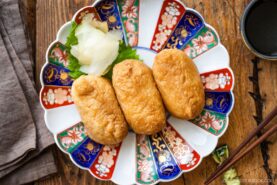 Three pieces of inari sushi on a decorative Japanese plate with colorful patterns, garnished with pickled ginger and a green leaf, placed on a wooden table with chopsticks and a small cup nearby.