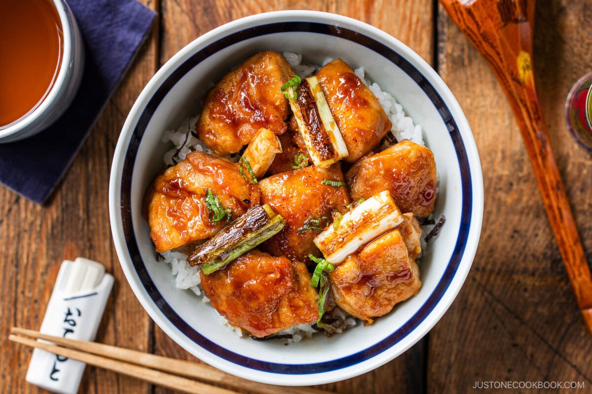 A bowl of rice topped with glazed grilled chicken pieces and scallions, this yakitori don is served with chopsticks on a wooden table. A cup and a sauce container are partially visible next to the bowl.