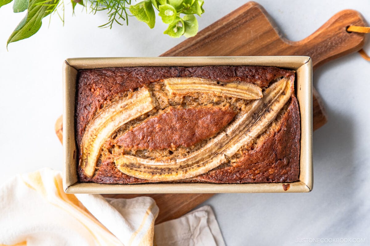 A loaf of Brown Butter Banana Bread in a rectangular baking pan, topped with two lengthwise banana slices, sits on a wooden cutting board with green plants and a yellow-striped towel nearby.