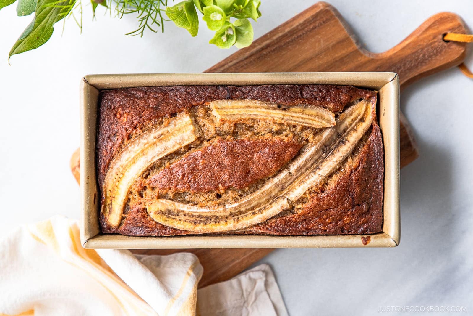 A loaf of Brown Butter Banana Bread in a rectangular baking pan, topped with two lengthwise banana slices, sits on a wooden cutting board with green plants and a yellow-striped towel nearby.