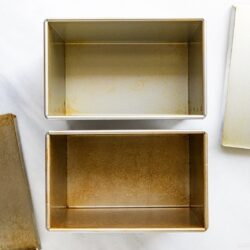 Two rectangular metal loaf pans with removable lids are shown open and empty on a white surface. The interior of each pan displays some discoloration or staining from use.
