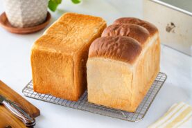 Two loaves of bread, one with a flat top and one with a rounded top, sit side by side on a cooling rack on a white surface. A knife, napkin, and potted plant are nearby.
