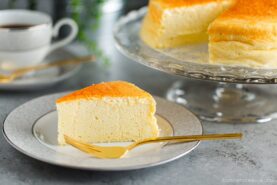 A slice of fluffy Japanese Cheesecake on a white plate with a gold fork, with the rest of the Japanese cheesecake recipe on a glass stand in the background. A cup of coffee and greenery are also visible.