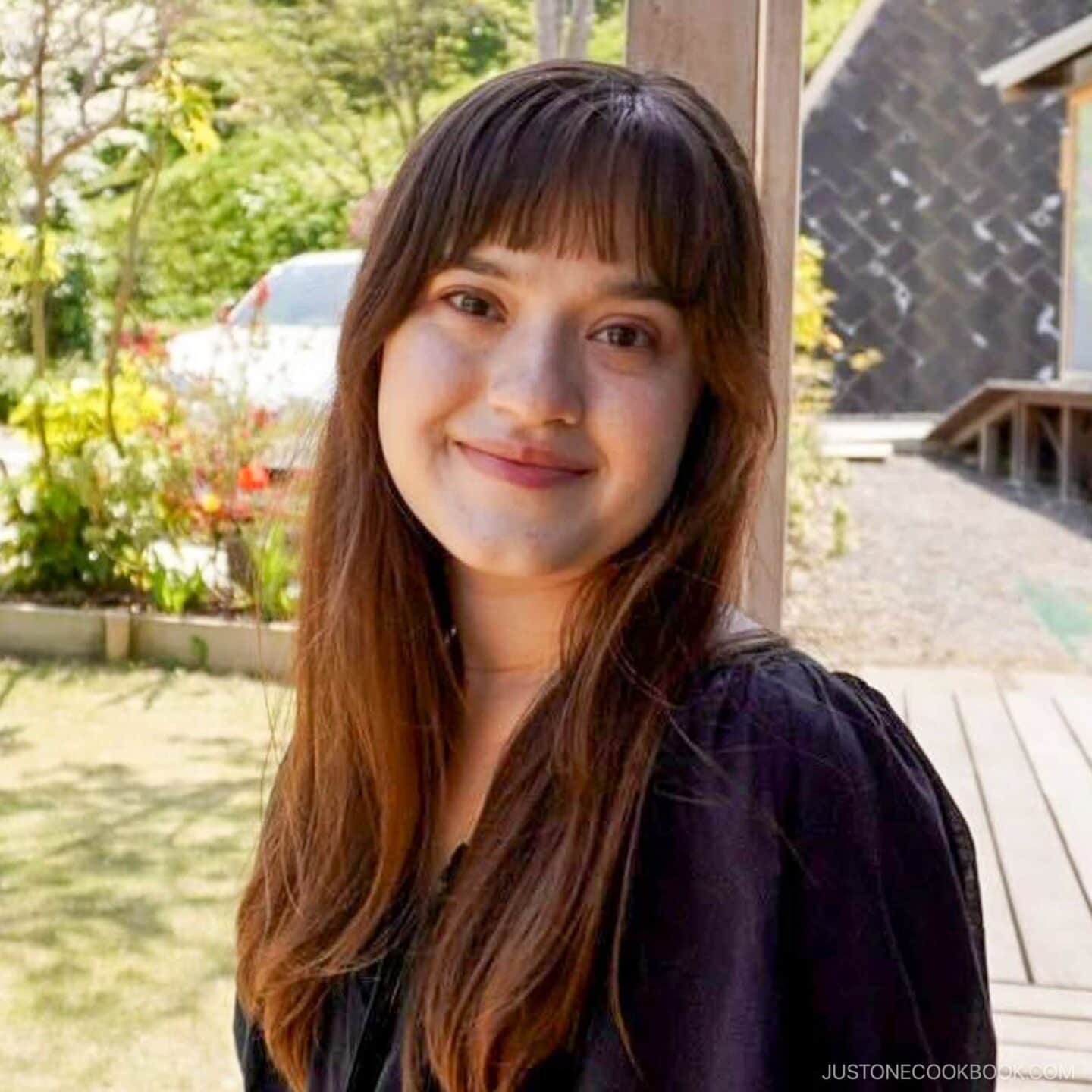 A young woman with long brown hair and bangs, wearing a black top, smiles while standing outdoors in a sunny garden with greenery and a building in the background.