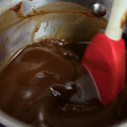 A red spatula stirs melted chocolate in a stainless steel pot, blending the smooth, glossy mixture along the sides of the pan, much like preparing a rich demi-glace recipe.