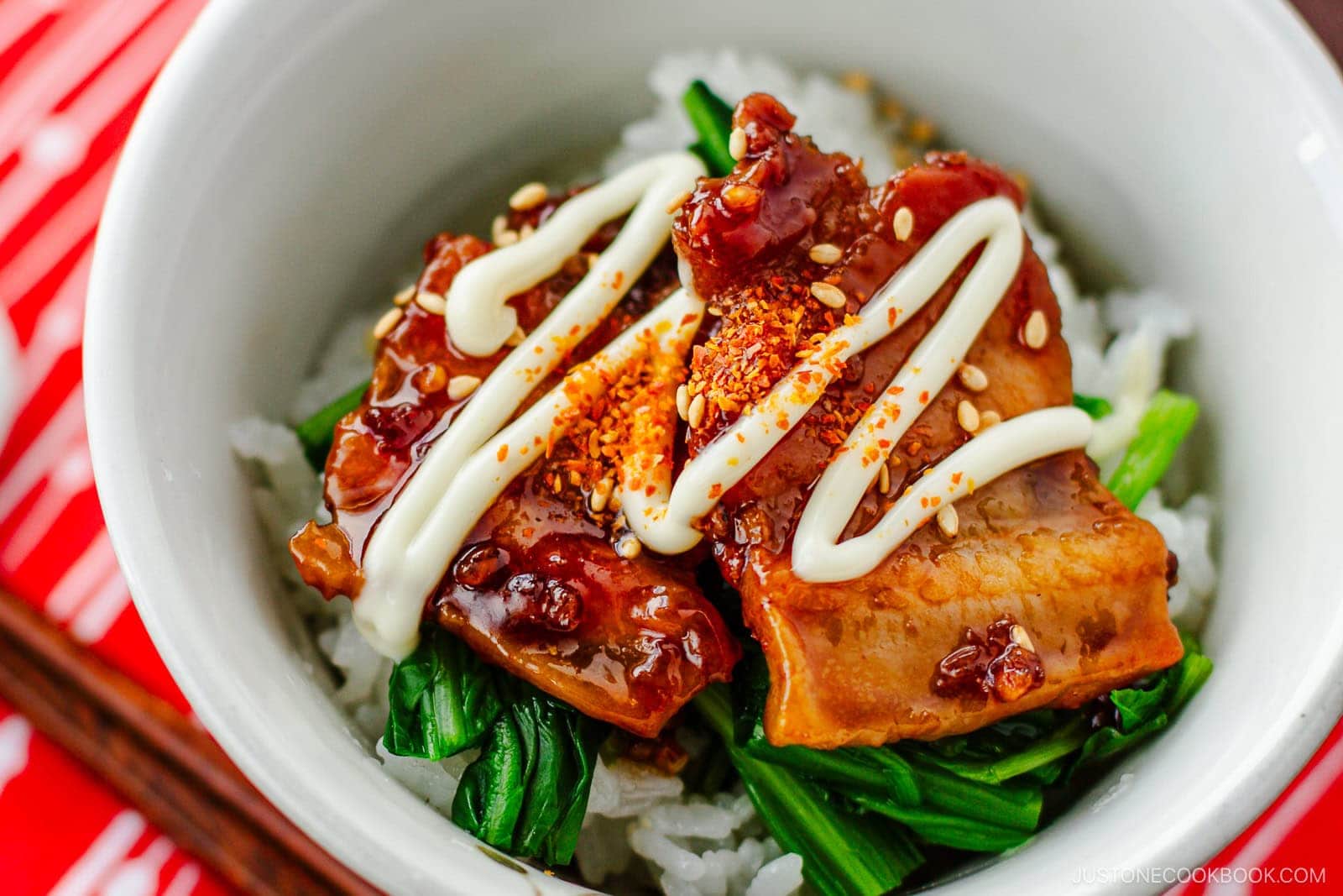 A bowl of rice topped with glazed grilled pork belly, Japanese mayonnaise, sesame seeds, chili flakes, and cooked greens, served with chopsticks on a red patterned mat.