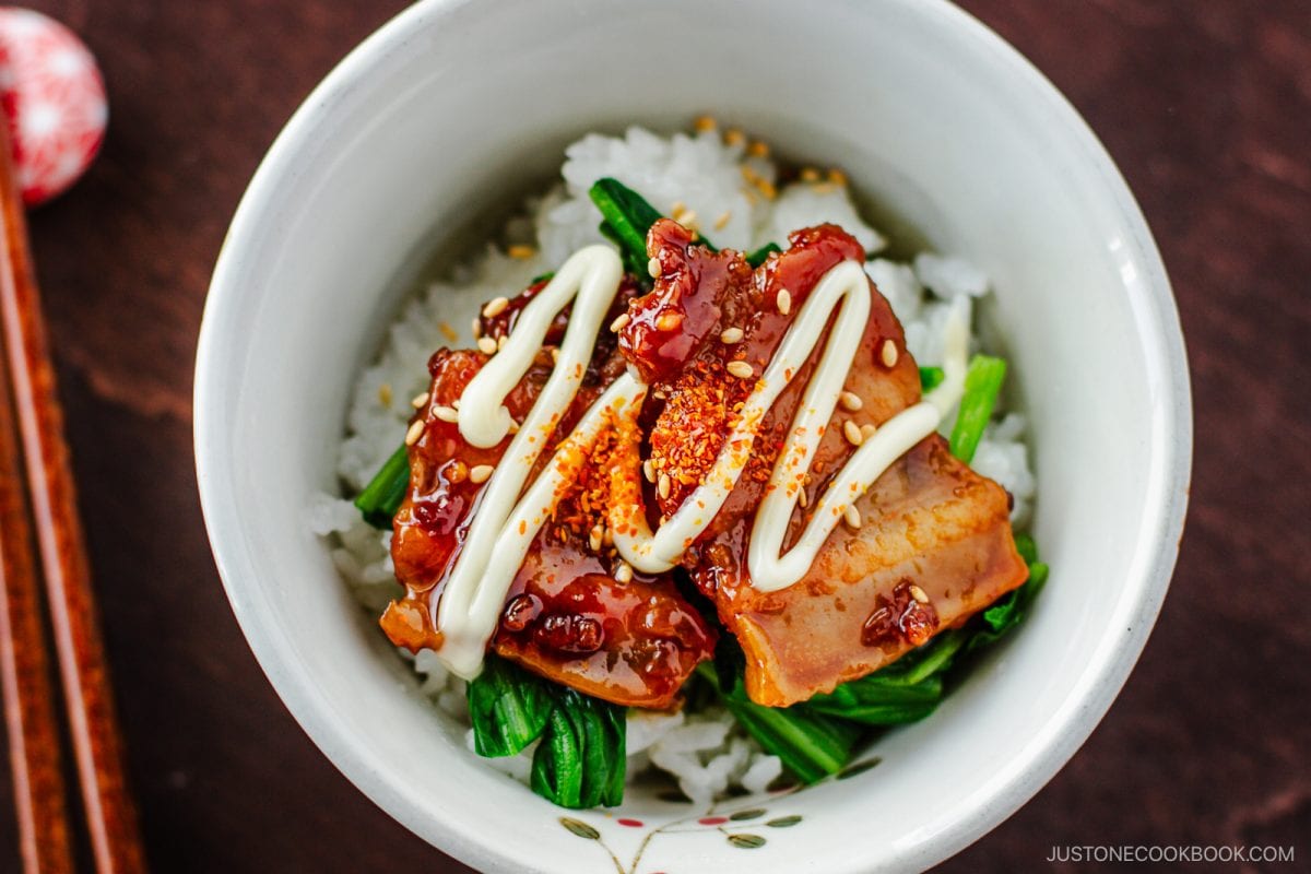 A bowl of white rice topped with glazed slices of meat, green vegetables, sesame seeds, and a drizzle of mayonnaise. Chopsticks are placed beside the bowl.
