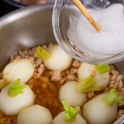 A hand pours a bowl of white liquid into a pot with small turnips, ground meat, and brown broth on the stovetop&mdash;demonstrating how to thicken sauce and soup with cornstarch slurry.
