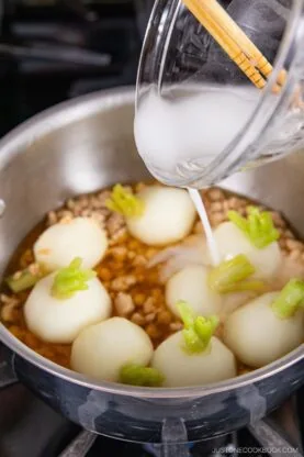 A hand pours a cloudy white liquid&mdash;like a cornstarch slurry used for how to thicken sauce and soup&mdash;from a measuring cup into a pot with whole turnips and chopped meat simmering in broth on the stovetop.