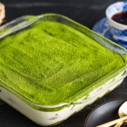 A glass dish filled with matcha tiramisu, topped with a smooth layer of vibrant green matcha powder, sits on a dark surface next to a blue patterned cup of coffee, gold fork and spoon, and a black plate.