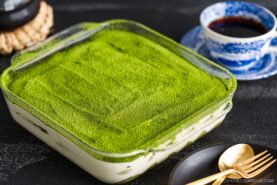 A glass dish filled with matcha tiramisu, topped with a smooth layer of vibrant green matcha powder, sits on a dark surface next to a blue patterned cup of coffee, gold fork and spoon, and a black plate.