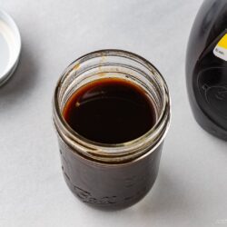 An open glass jar filled with dark yakisoba sauce, made from a classic yakisoba sauce recipe, sits on a white surface next to its lid and a bottle labeled &ldquo;Yakisoba Sauce.&rdquo;.