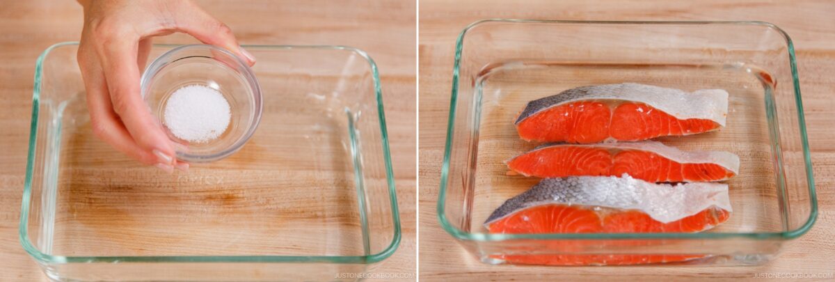 A hand pours salt from a small glass bowl into a glass baking dish; beside it, three raw salmon fillets with skin on are arranged for making Kobumaki (Salmon Kombu Roll) on a wooden surface.
