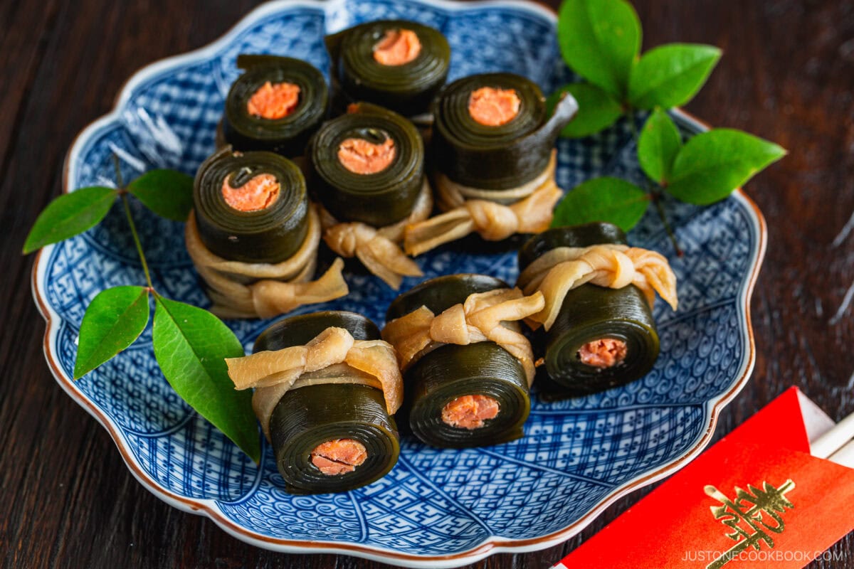 A blue patterned plate holds neatly arranged Kobumaki (Salmon Kombu Roll)&mdash;kelp seaweed rolls with salmon filling, tied with a light-colored gourd strip and garnished with green leaves. Red envelope chopsticks and a napkin are beside the plate.