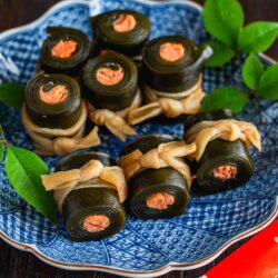 A blue patterned plate holds neatly arranged Kobumaki (Salmon Kombu Roll)—kelp seaweed rolls with salmon filling, tied with a light-colored gourd strip and garnished with green leaves. Red envelope chopsticks and a napkin are beside the plate.