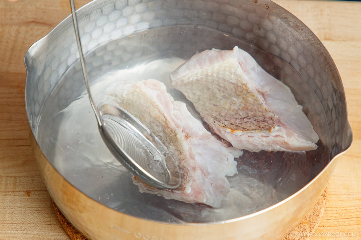 Two raw fish fillets are undergoing prep work as they&rsquo;re blanched in a pot of hot water, with a metal skimmer partially submerged beside them. The pot rests on a wooden surface.