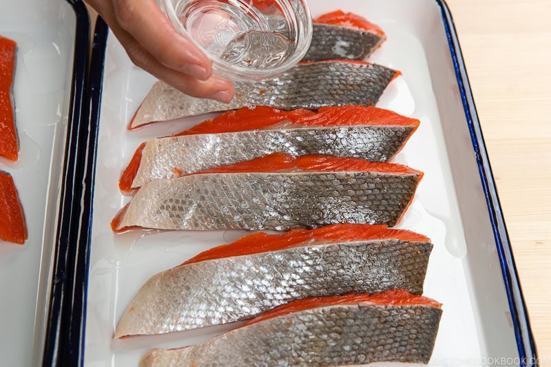 A hand performs prep work, pouring water from a small glass bowl over several raw salmon fillets with shiny skin, arranged neatly on a white tray.