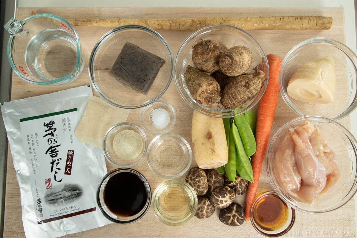 A top-down view of various ingredients for Instant Pot Nishime, including chicken, root vegetables, shiitake mushrooms, snow peas, konnyaku, soy sauce, mirin, dashi packet, and seasonings on a wooden surface.