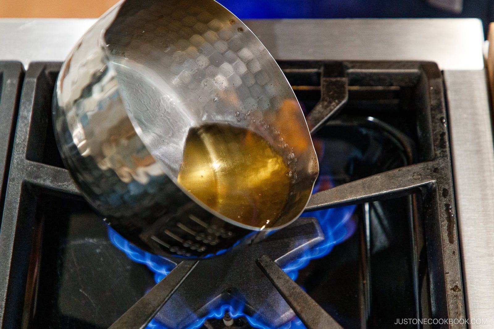 A metal pot with oil and nikiri mirin is tilted over a blue gas flame on a stove burner.