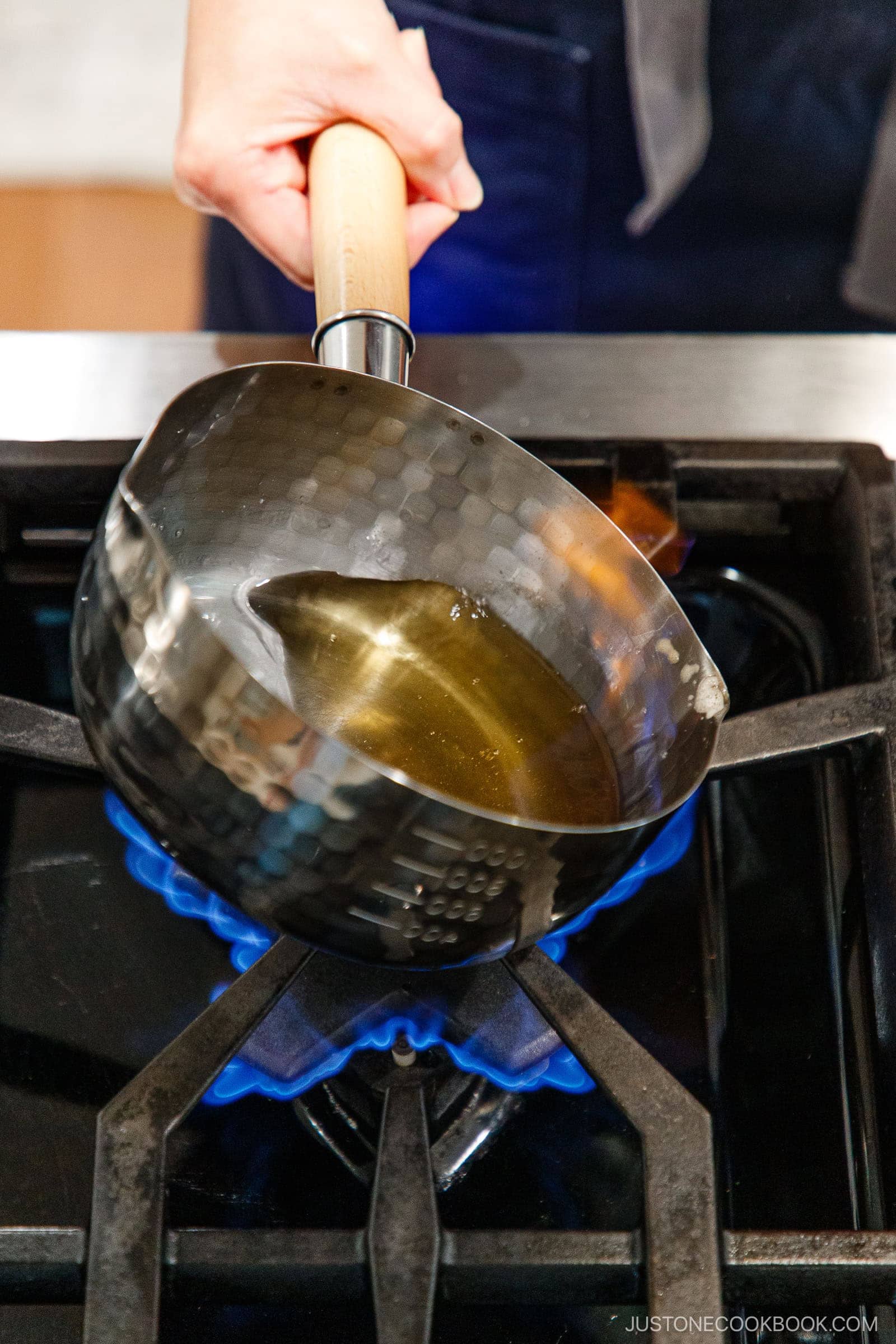 A hand holds a stainless-steel saucepan with oil and a splash of nikiri mirin over a blue gas flame on a stovetop, heating the mixture.