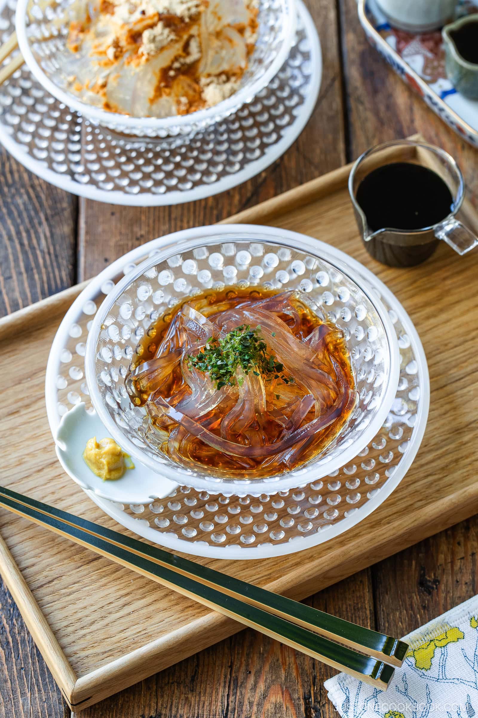A clear bowl of Japanese kanten noodles with sauce and garnish sits on a wooden tray with chopsticks, a dollop of mustard, and a small pitcher of dark sauce beside it. Another dessert is partly visible in the background.