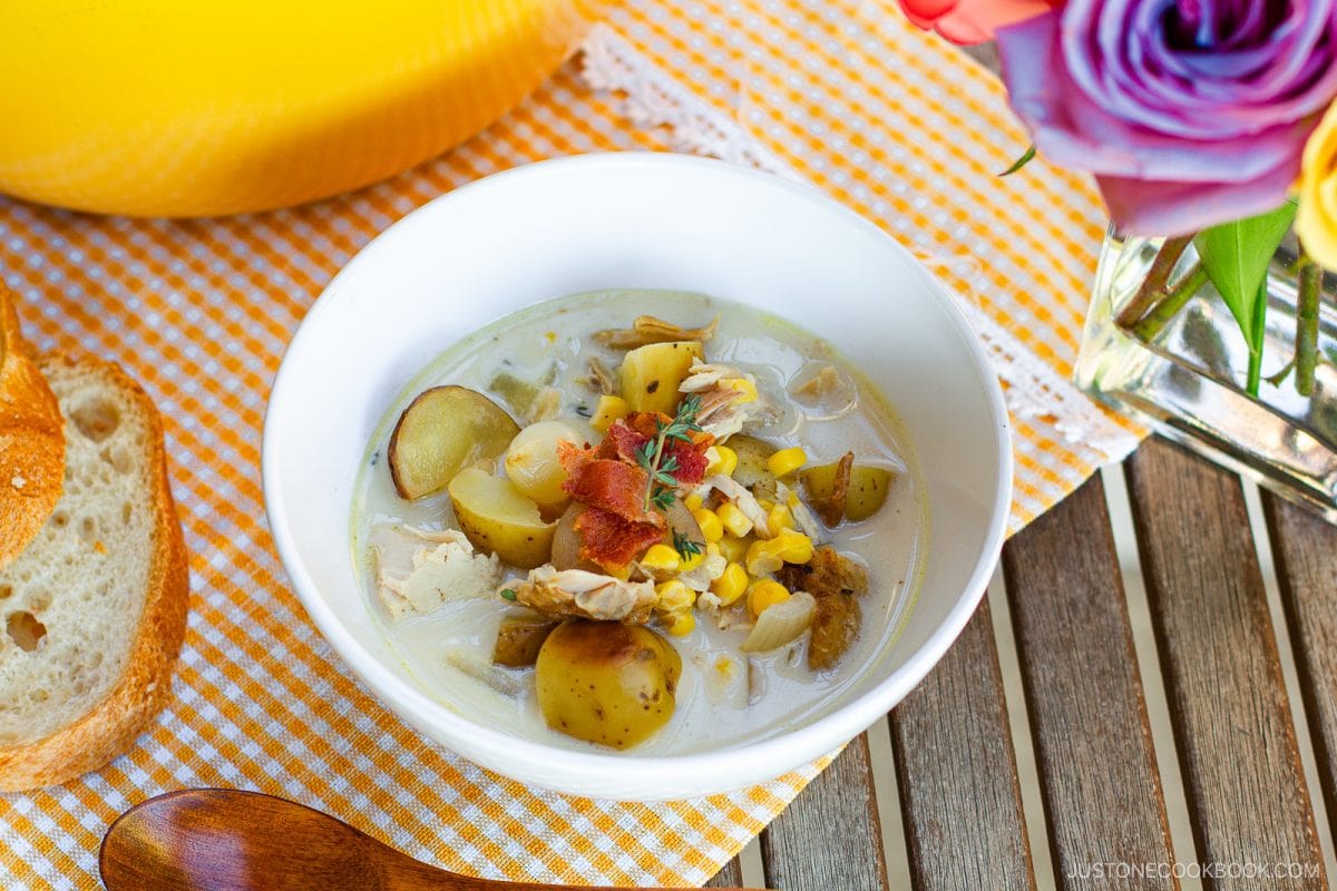 A white bowl of creamy Chicken Corn Chowder, brimming with tender chicken, potatoes, corn, and crispy bacon, sits on a checkered cloth next to sliced bread, a yellow pot, and a vase of colorful flowers.