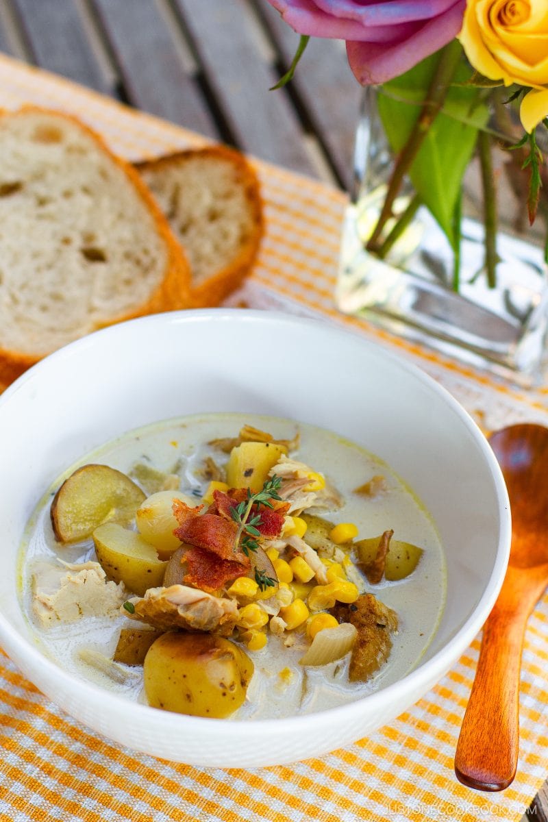 A bowl of creamy Chicken Corn Chowder with potatoes, corn, chicken, and bacon, garnished with herbs. Two slices of bread, a wooden spoon, and a vase of colorful flowers are nearby on a checkered cloth.
