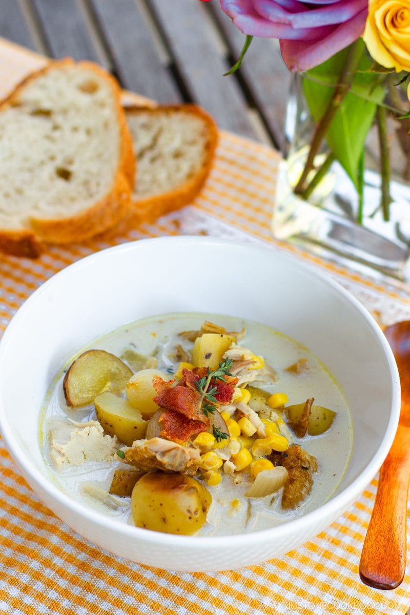 A bowl of creamy Chicken Corn Chowder with tender chicken, potatoes, sweet corn, bacon, and fresh herbs sits on a checkered cloth beside slices of bread and a vase of purple and yellow flowers.