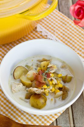 A white bowl of creamy Chicken Corn Chowder with potatoes, topped with bacon and herbs, sits on a yellow checkered cloth near a yellow pot and a pink rose.