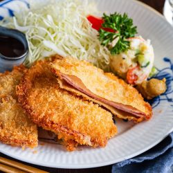 A plate with breaded, fried pork cutlets&mdash;reminiscent of Ham Katsu from Midnight Diner&mdash;shredded cabbage, potato salad, a dollop of mustard, and a small dish of dark sauce. Chopsticks and a drink are beside the plate.