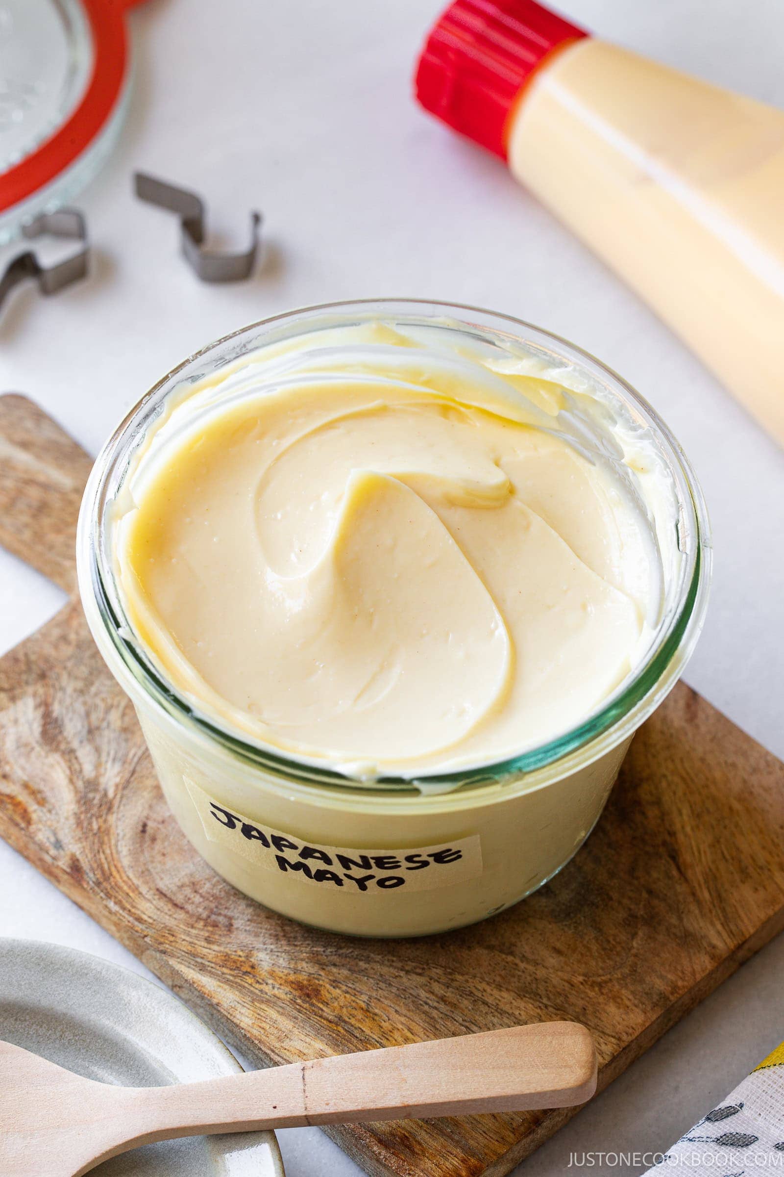 A glass jar labeled Japanese Mayo filled with creamy mayonnaise sits on a wooden board, with a tube of mayo and kitchen utensils nearby.