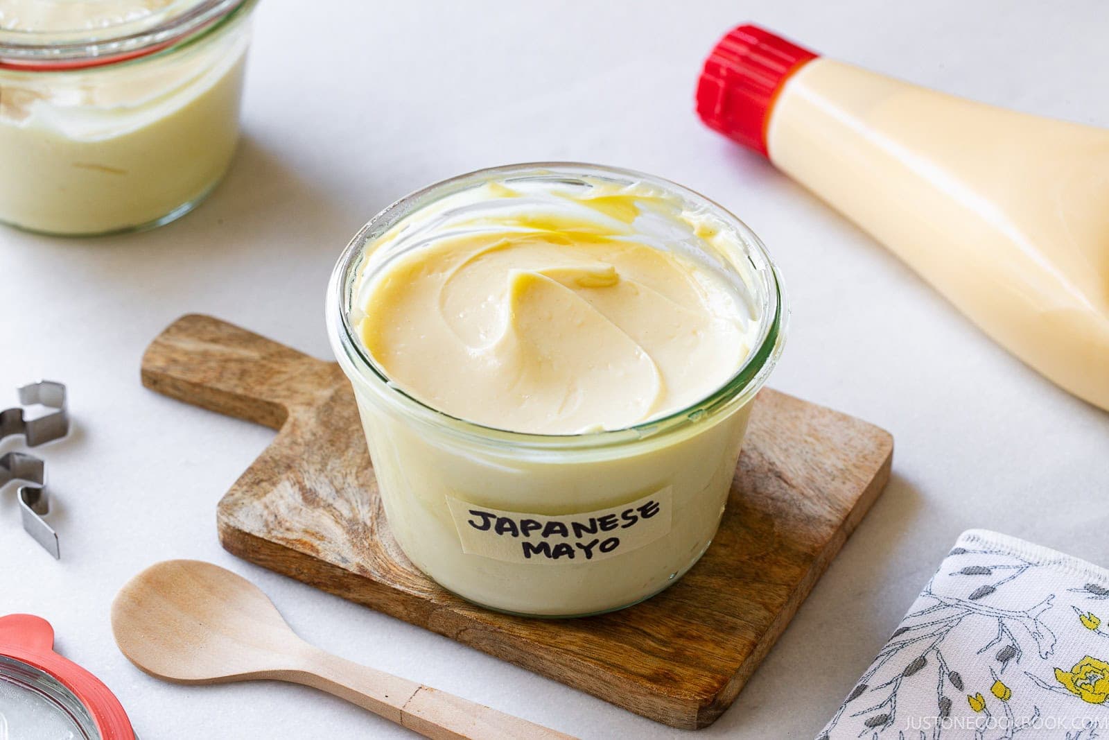 A glass jar labeled Japanese Mayo sits on a small wooden board. Nearby are a wooden spoon, a squeeze bottle, a fabric napkin, and another jar of mayonnaise. The setting is bright and clean.