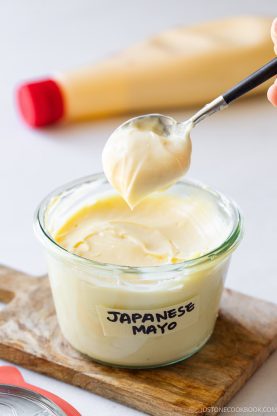 A spoon scooping creamy Japanese mayo from a glass jar labeled Japanese Mayo, with a squeeze bottle in the background on a wooden board.