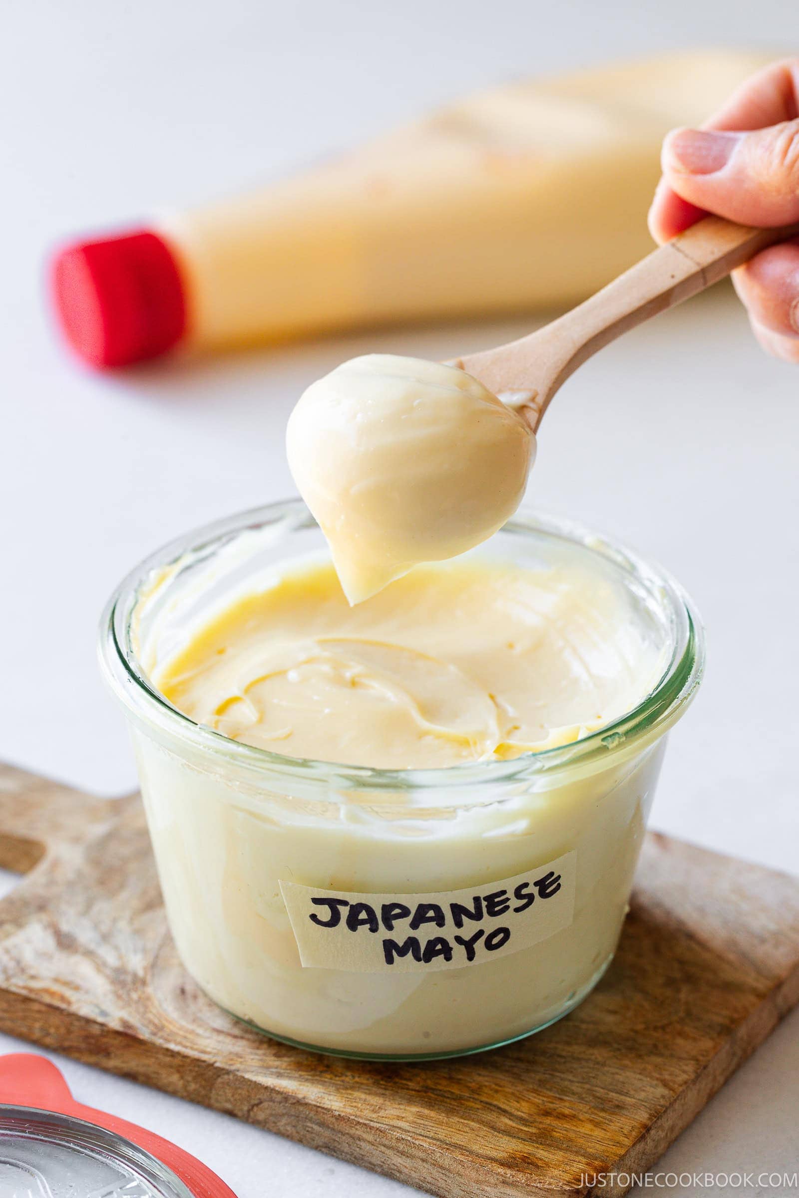 A wooden spoon scoops creamy Japanese mayo from a clear glass jar labeled Japanese Mayo, with a squeeze bottle and lid nearby on a wooden board.