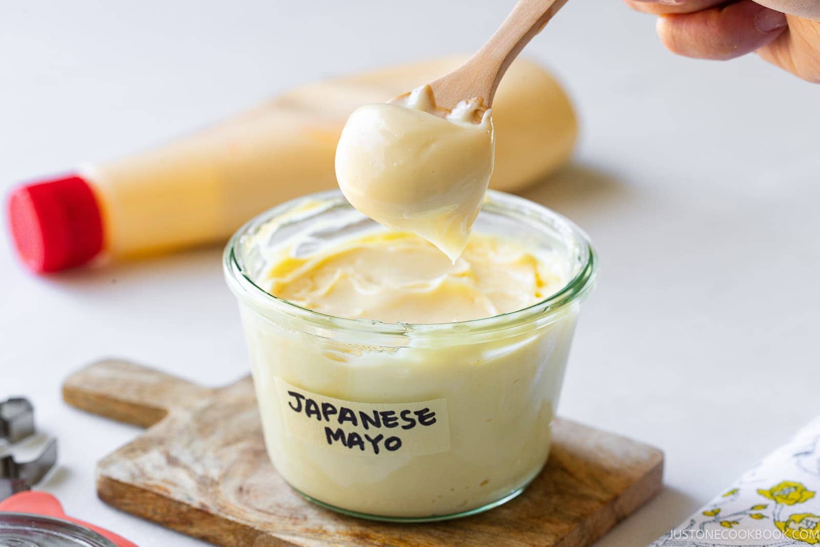 A glass jar labeled Japanese Mayo sits on a wooden board. A hand holds a small spoon with mayo above the jar. A squeezable bottle with a red cap is in the background.