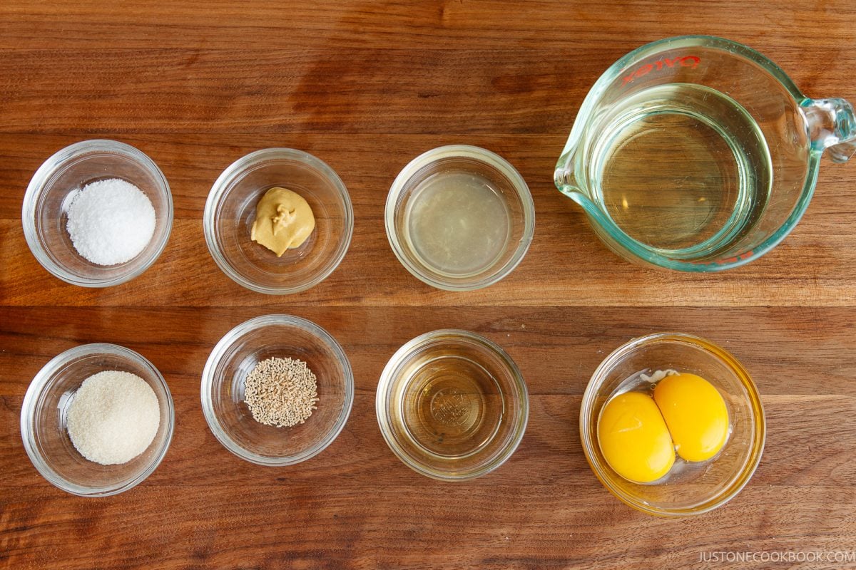 Eight small glass bowls and one large measuring cup contain ingredients on a wooden surface, including oil, salt, mustard, vinegar, sugar, sesame seeds, seasoning, and two egg yolks.