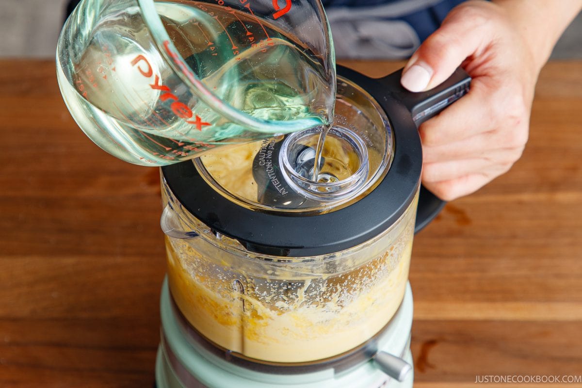 A person pours oil from a measuring cup into a food processor blending japanese mayonnaise mixture, on a wooden surface.