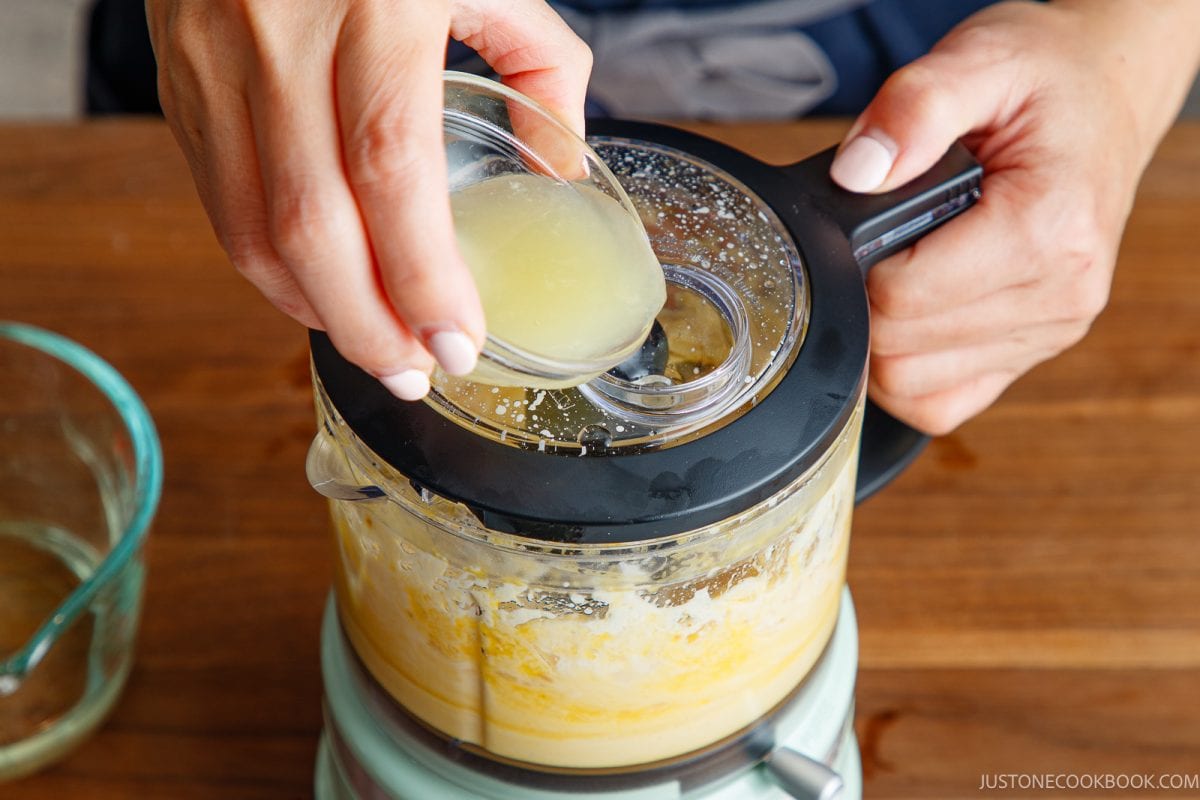 A person pours lemon juice from a small glass bowl into a food processor blending japanese mayonnaise mixture, on a wooden countertop.