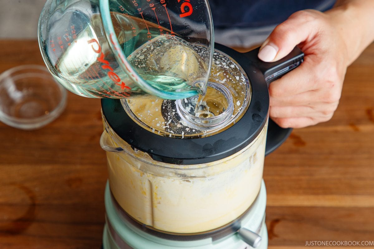 A person pours oil from a measuring cup into a blender filled with a creamy japanese mayonnaise mixture on a wooden surface.