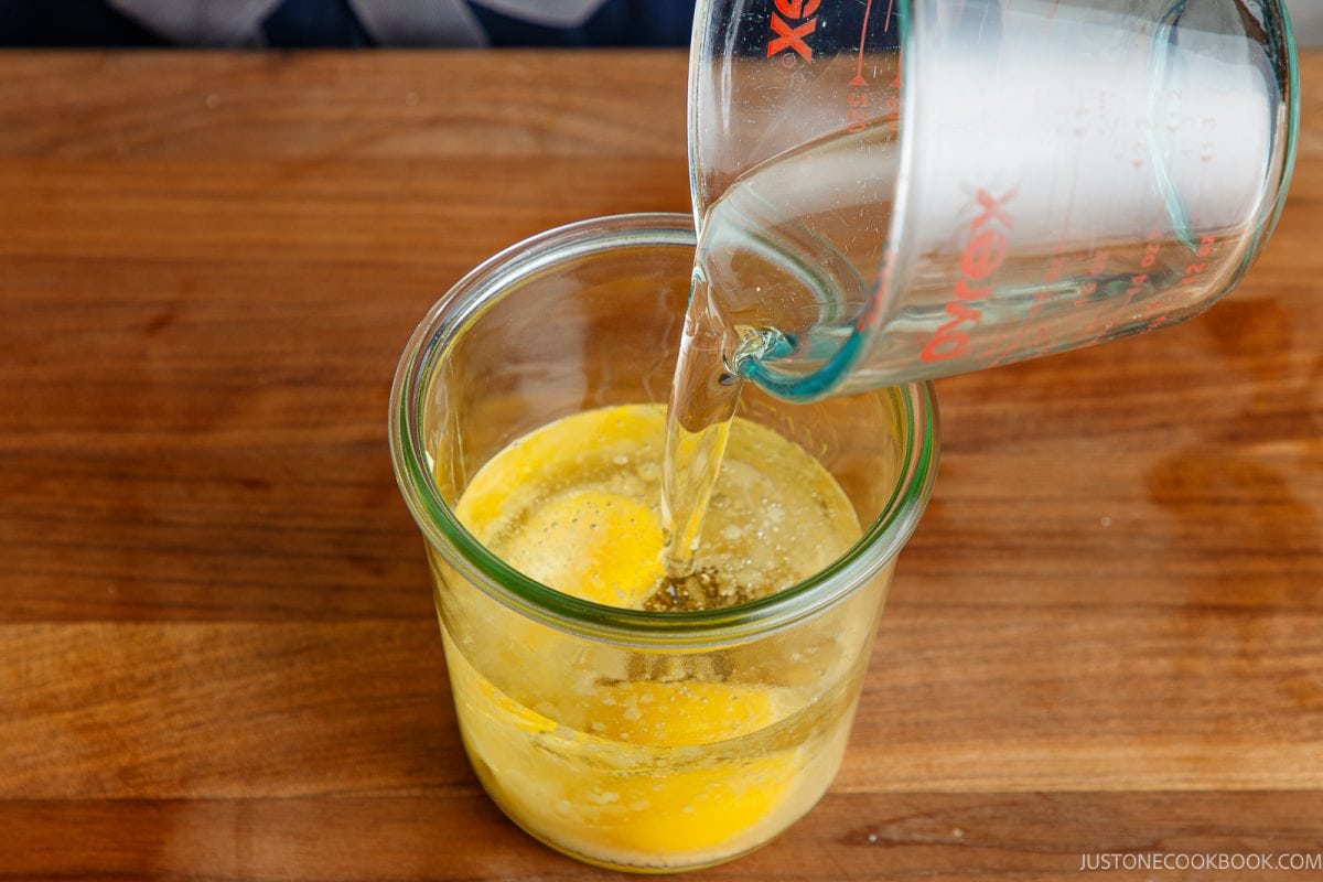 A glass measuring cup pours oil into a glass bowl containing two cracked eggs and a dollop of Japanese mayonnaise on a wooden surface.