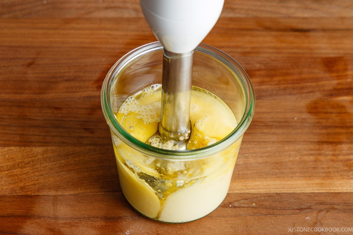 A hand blender is blending eggs, oil, and other ingredients in a glass jar on a wooden surface to make homemade Japanese mayonnaise.