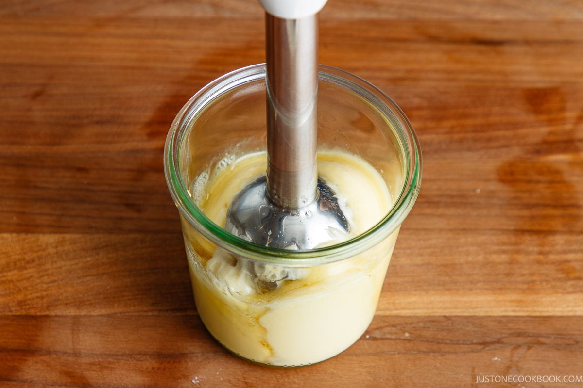 An immersion blender is mixing creamy Japanese mayonnaise ingredients in a glass jar on a wooden surface.
