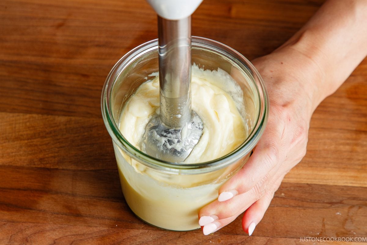 A hand holds a glass jar while using an immersion blender to mix a creamy homemade Japanese mayonnaise&mdash;on a wooden surface.