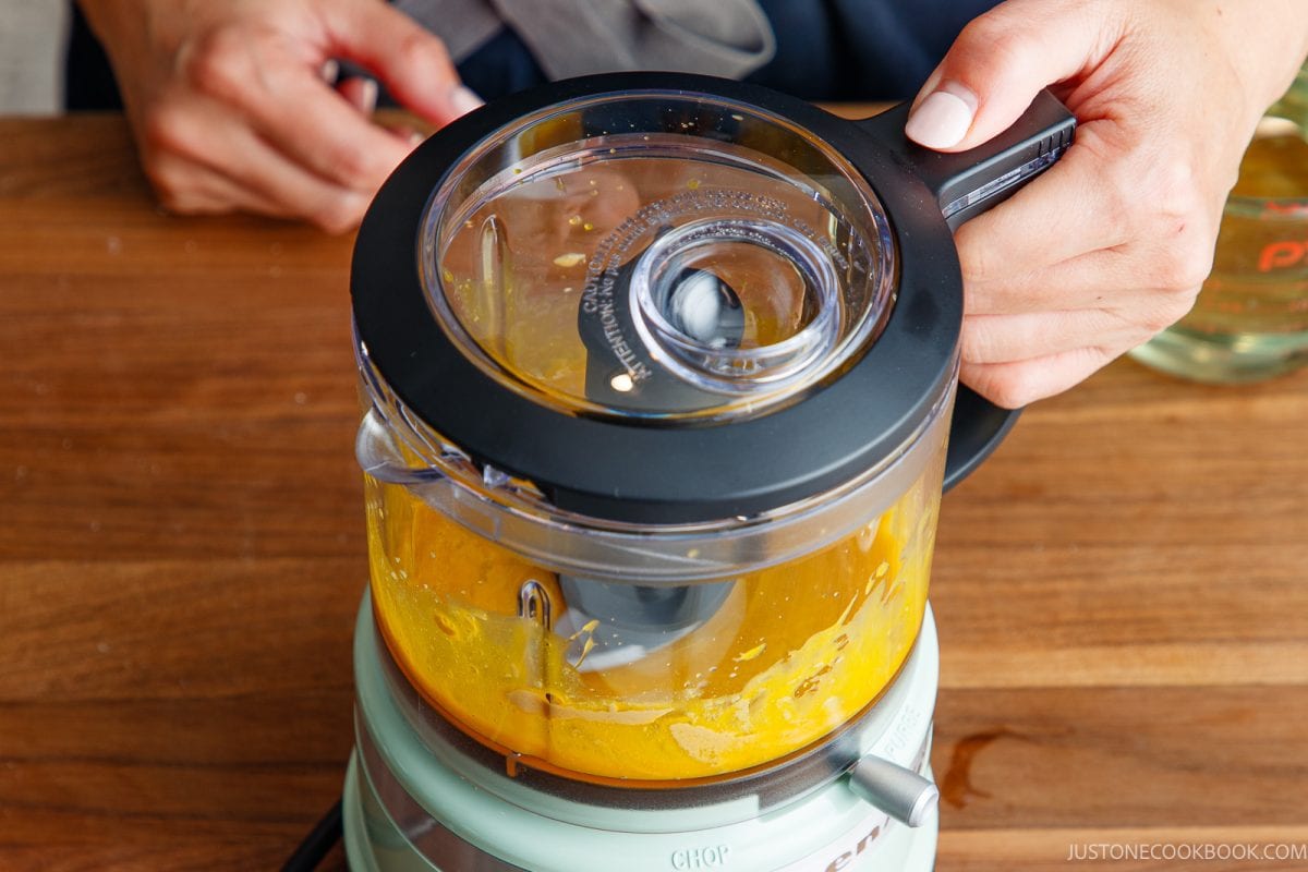 Close-up of hands using a food processor to blend Japanese mayonnaise on a wooden table. The lid is partially transparent, showing the creamy contents inside.