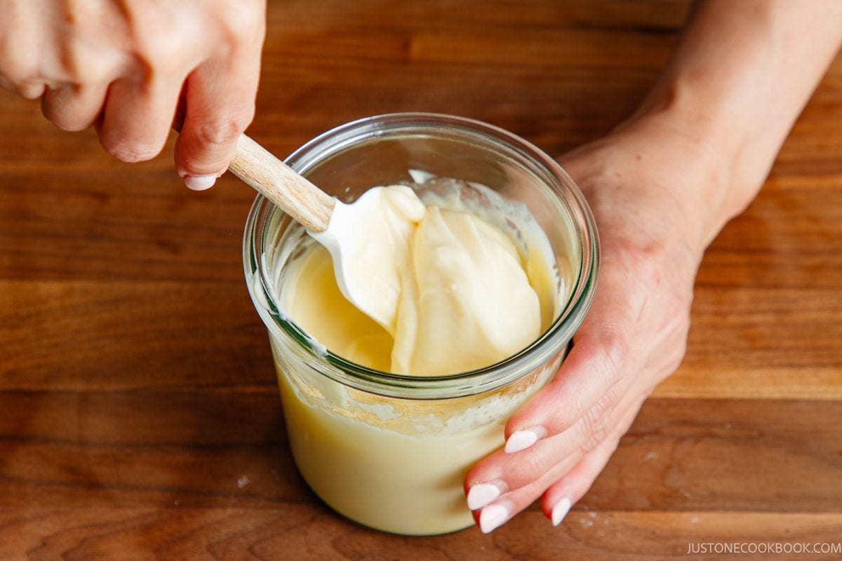 A person stirs Japanese mayonnaise, in a glass jar with a white spatula on a wooden surface.