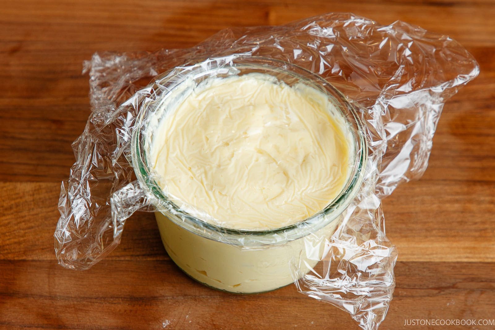 A glass container filled with a creamy, pale yellow substance&mdash;reminiscent of smooth Japanese mayonnaise&mdash;partially covered with clear plastic wrap, sits on a wooden surface.
